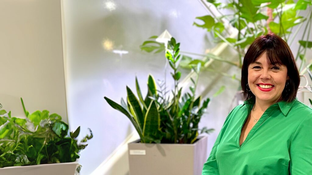 Minna Harju istumassa viherkasvien edessä Cursorin aulassa. Hänellä on päällään kirkkaan vihreä paita.